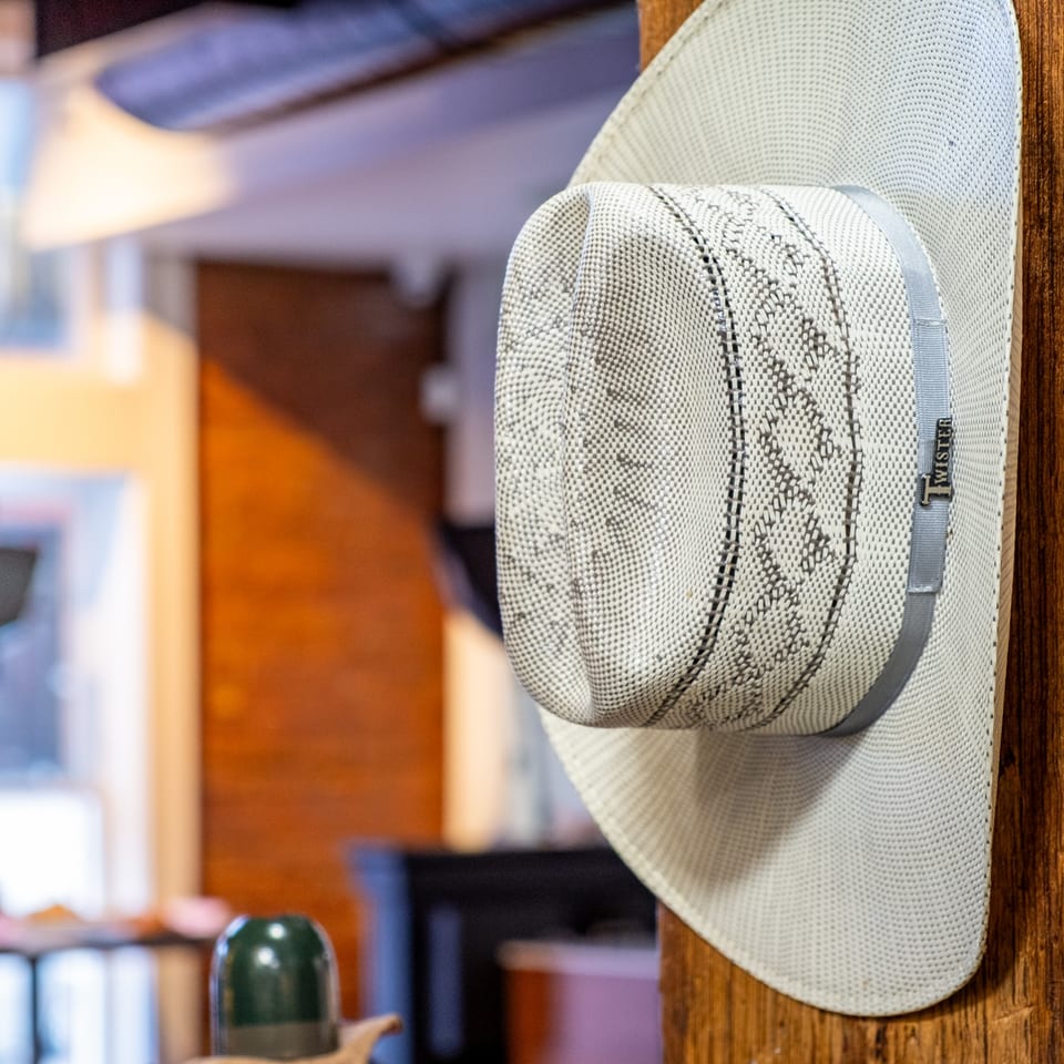 A light-colored woven cowboy hat with a gray band hangs on a wooden post.