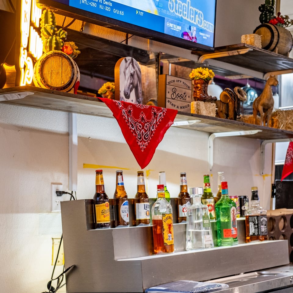 A bar shelf with various liquor bottles, beer, and Western-themed decor under a TV screen.
