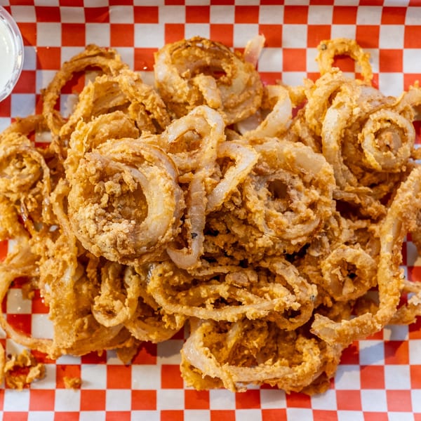 A large serving of crispy, golden-brown onion rings on red and white checkered paper with ranch.
